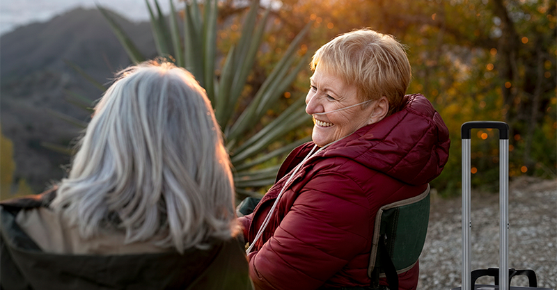 2 old women sitting outside in camping chairs smiling and one of them is using the Kingon P2-TOC transportable oxygen concentrator.