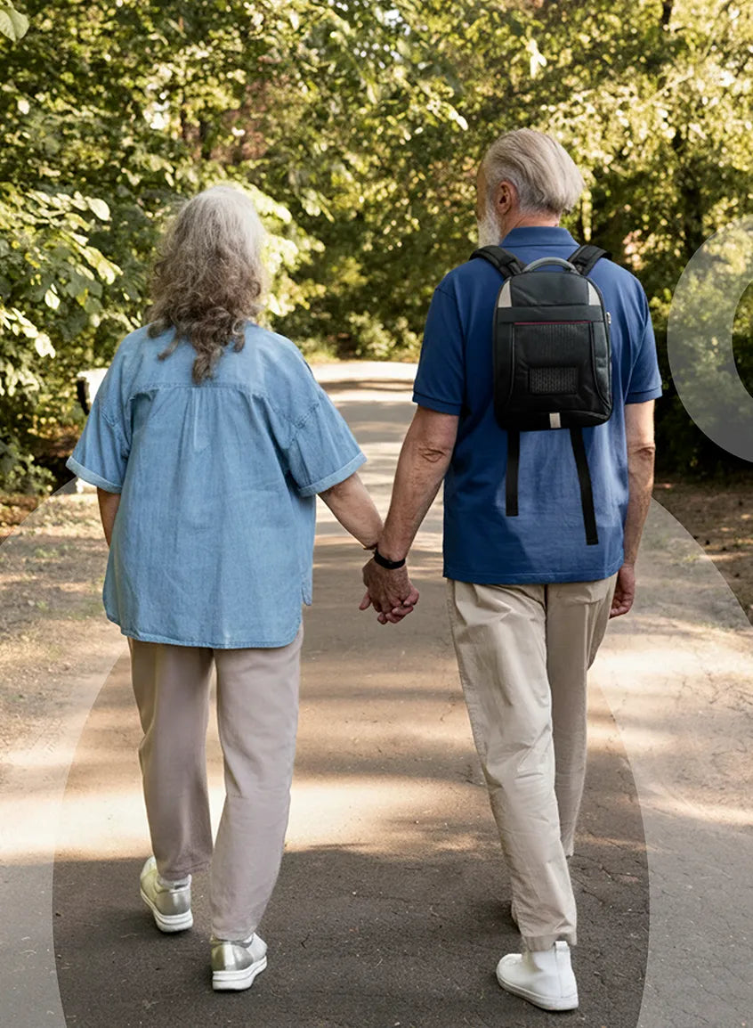 Two elderly people walking hand in hand on a path with trees in the background. The man has a Kingon backpack on.