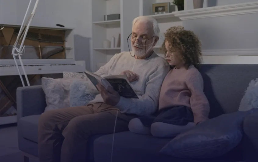 Grandpa and grandchild sitting on a couch reading a book and grandpa is using a nasal cannula.