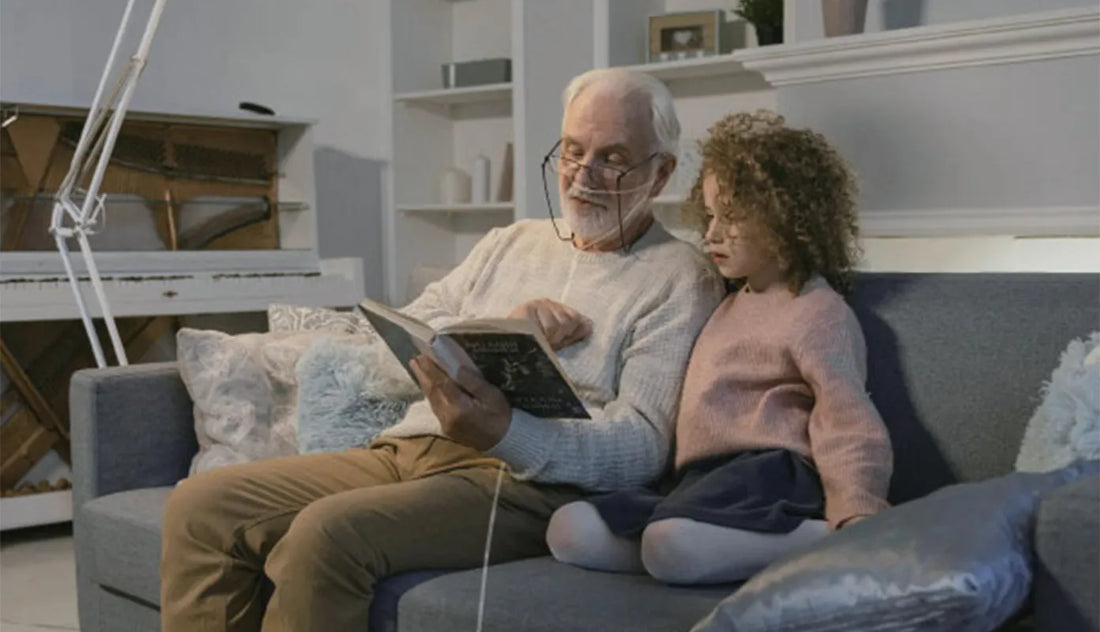 Grandpa and grandchild reading a book on a couch. Grandpa has a nasal cannula on.