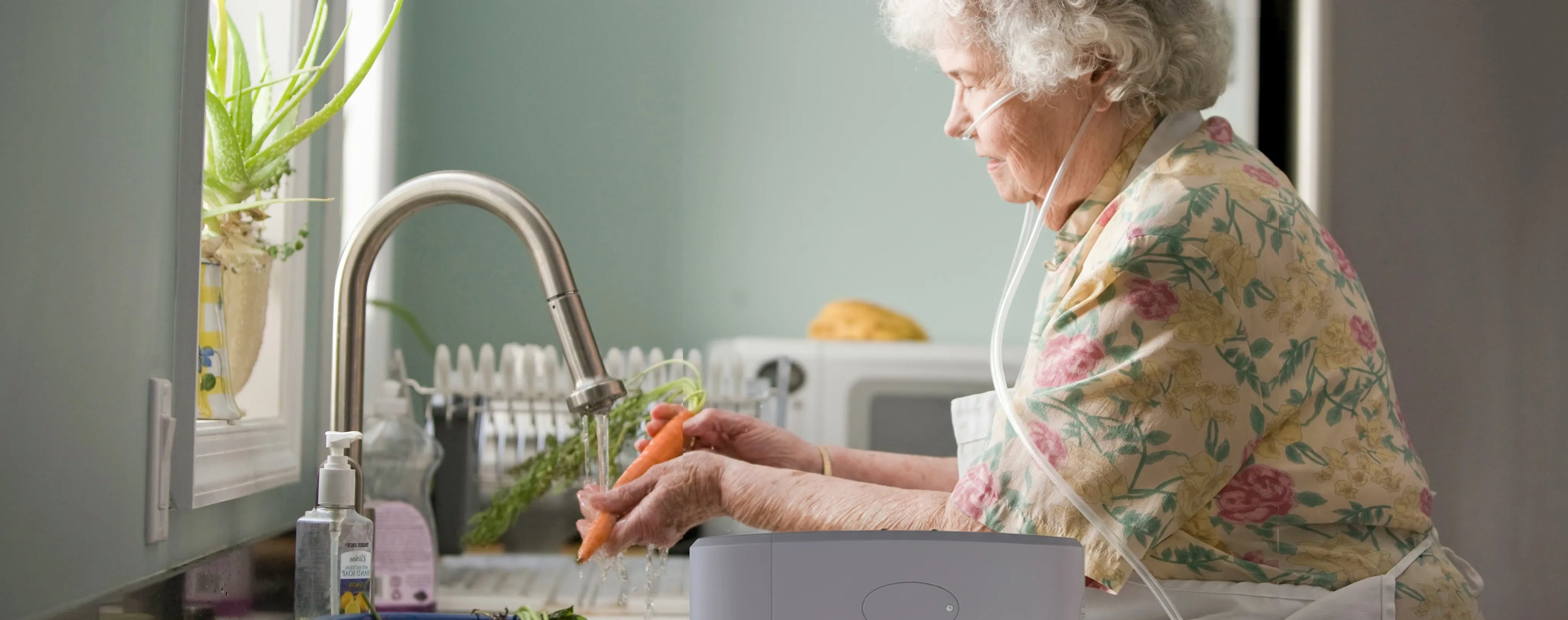 Old woman at home washing a carrot while using a Kingon portable oxygen concentrator.