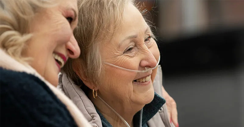 Daughter and old mother smiling and hugging while looking into the distance. Mother has a nasal cannula on.
