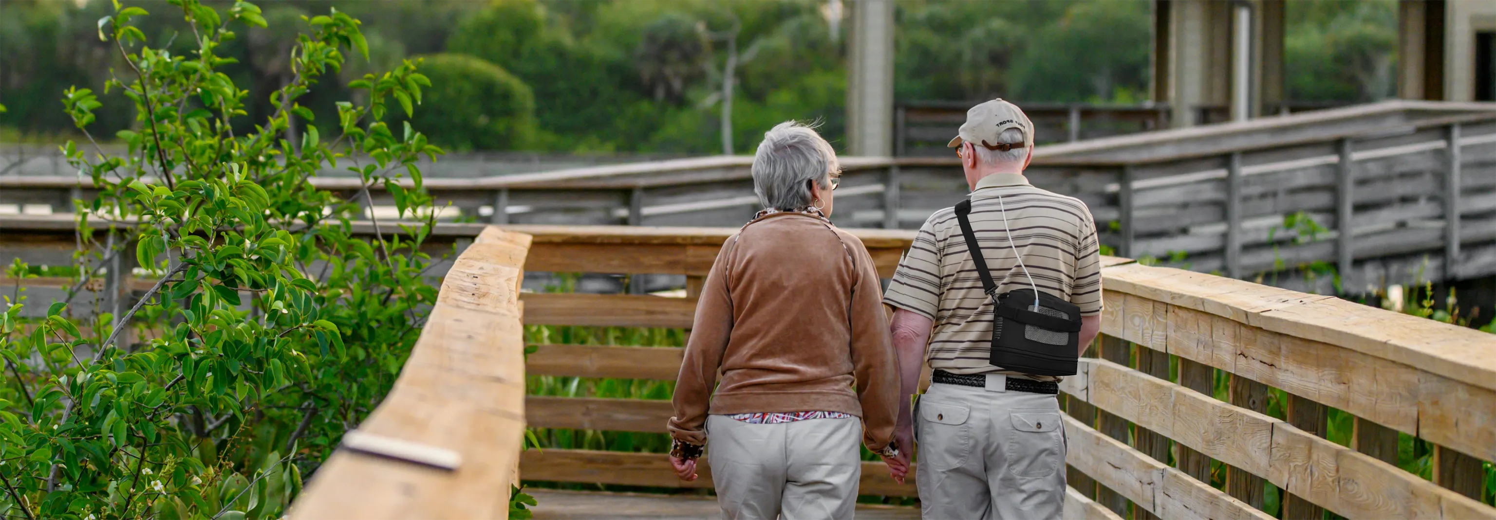 Old couple walking on a deck and the old man has a Kingon P2 portable oxygen concentrator on with the shoulder carry bag.