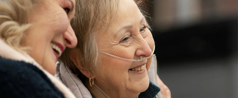Two elderly women smiling, one with a visible nasal cannula.